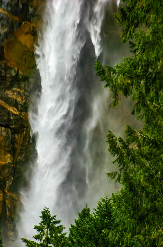 Close-up view of a powerful waterfall cascading down a rocky cliff, partially framed by lush green evergreen trees.
