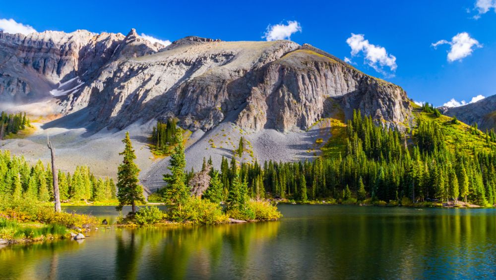 Mountain lake scene with evergreen trees reflecting in the calm water, under a bright blue sky with rugged rocky peaks in the background.