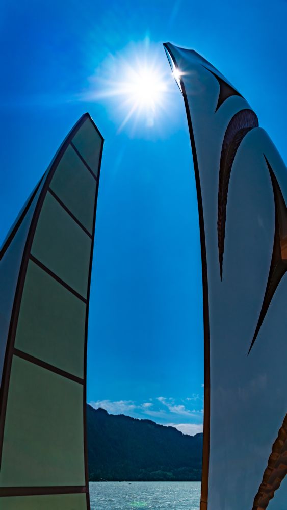 Two tall, curved Indigenous art sculptures frame the sun high in a clear blue sky, with a shimmering lake and mountain silhouette below.