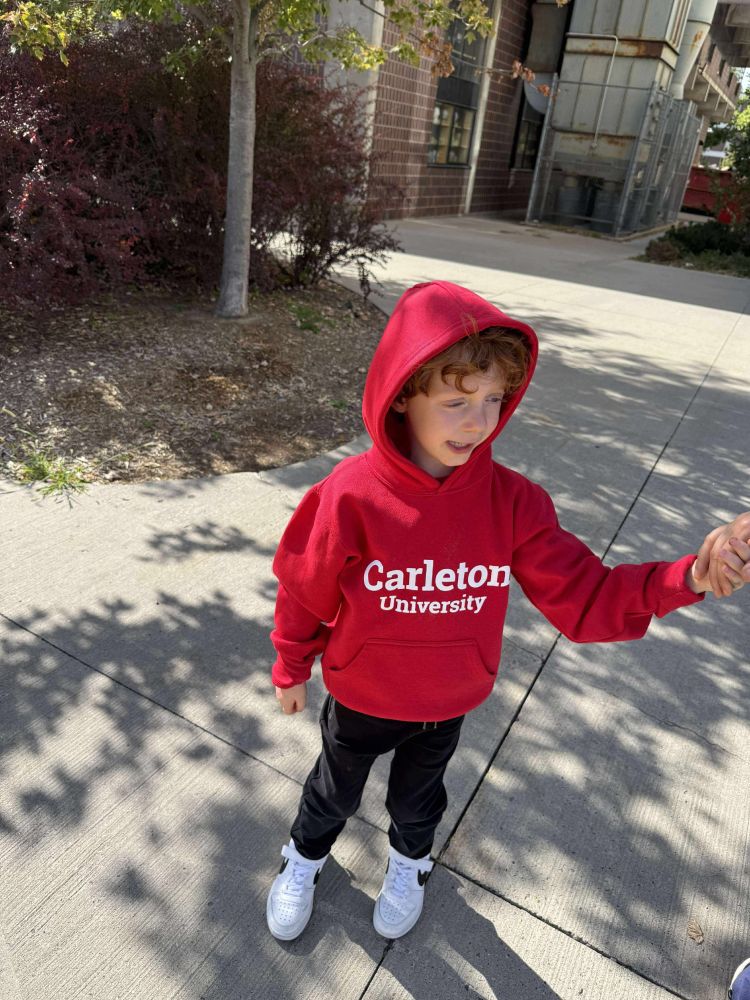 A boy stands outside in a red hoodie and black pants and white shoes. 