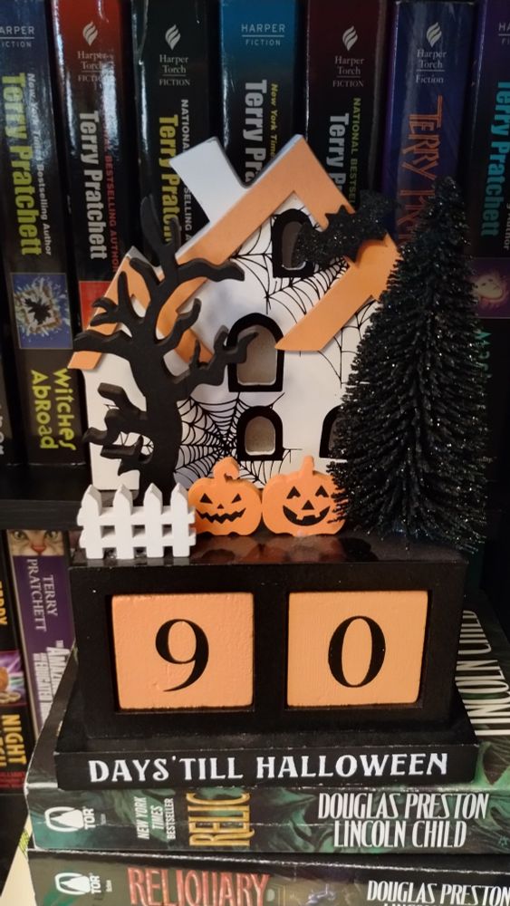 Photo of a decorative wooden countdown calendar for Halloween. Two large orange cubes at the bottom together form "90" above the words "Days 'till Halloween". Above is a white haunted house with orange roof snd black window trim. The house is decorated in cobwebs, with a pair of jack-o-lanterns and black trees out front. Ironically, several books by Terry Pratchett are in thr background, with the title "Witches Abroad" displayed immediately next to the house.