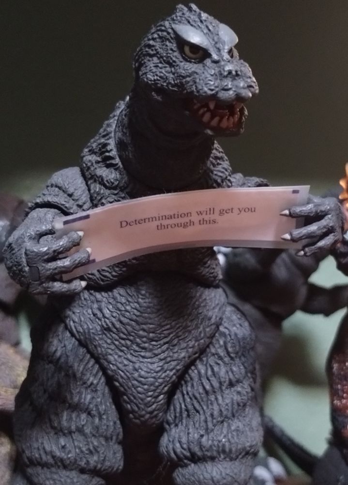 Close-up photo of a detailed toy Godzilla figure holding a laminated fortune cookie fortune in his hands. It reads "Determination will get you through this."