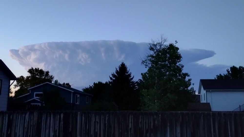 A hazy grey-blue sky over a suburban landscape. The sky is perfectly clear except for a huge, flat-topped anvil-shaped thunderhead looming ominously in the distance.
