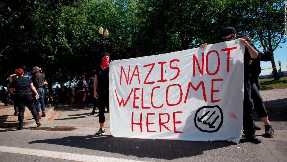 drei vermummte menschen halten ein transparent mit dem spruch: "nazis not welcome here" und dem logo der eisernen front (drei schräg nach unten links weisende pfeile in einem kreis)