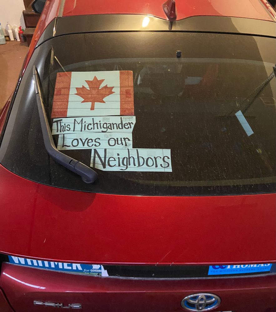 Freehand Canadian flag drawing with the text "This Michigander Loves our Neighbors" taped to the inside of the hatchback window of a red car.
