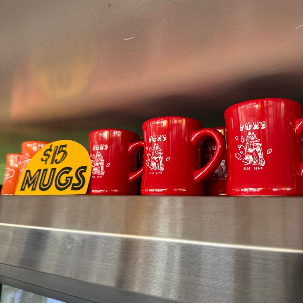 A refrigerator topped with a row of Fox’s mugs, which are presented next to a small sign that says, “$15 Mugs.” There are two Cindy’s mugs displayed behind the small sign. 