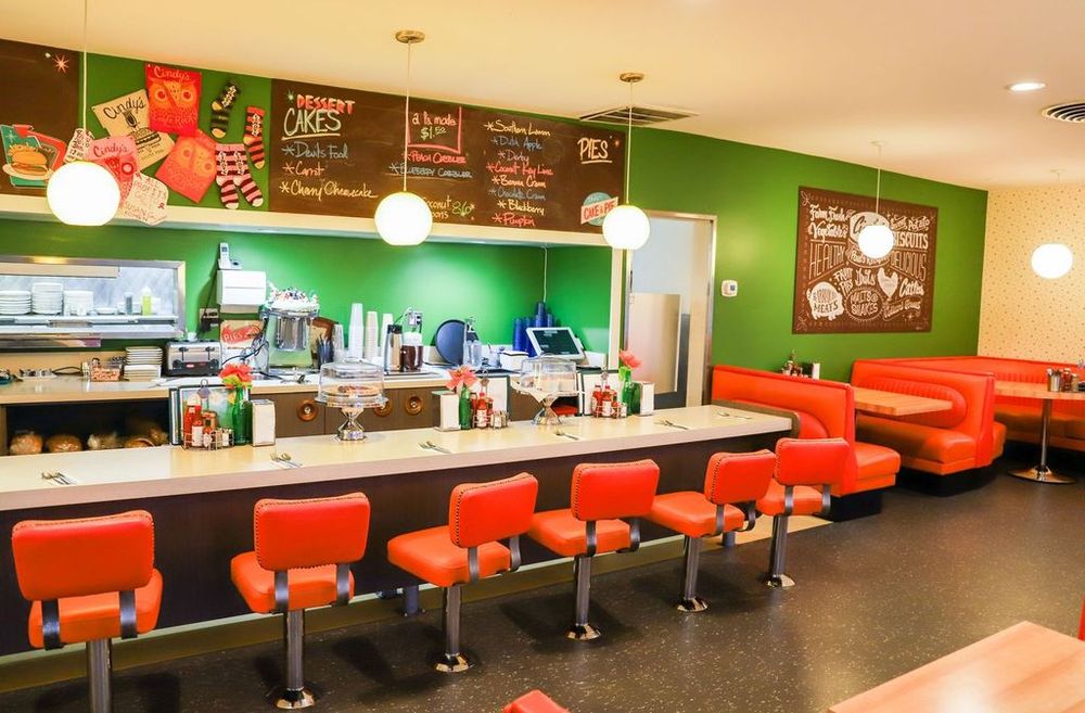 The interior of Cindy's, featuring a counter space for dining with vibrant orange chairs, matching orange corner booths, retro-inspired pendant lights, quirky merchandise pinned to the walls, a colorful chalkboard dessert menu, charming wall art, and a small open-window view of the kitchen area.