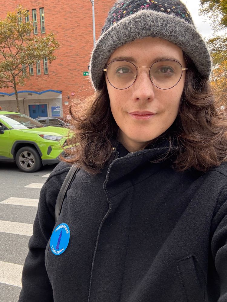 Eileen smiling in front of a NYC crosswalk in a black coat with a winter hat and glasses, and a round blue sticker that says “Manhattan Votes Early” with a little Empire State Building in the middle of the sticker.