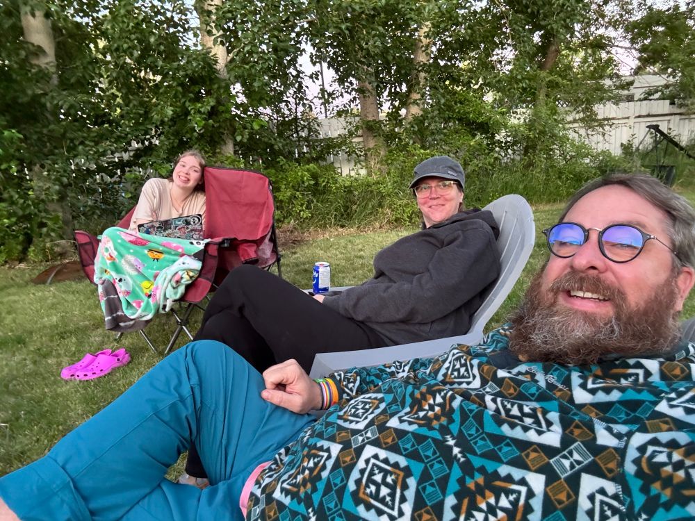 Three people in lawn chairs, sitting in the backyard. 