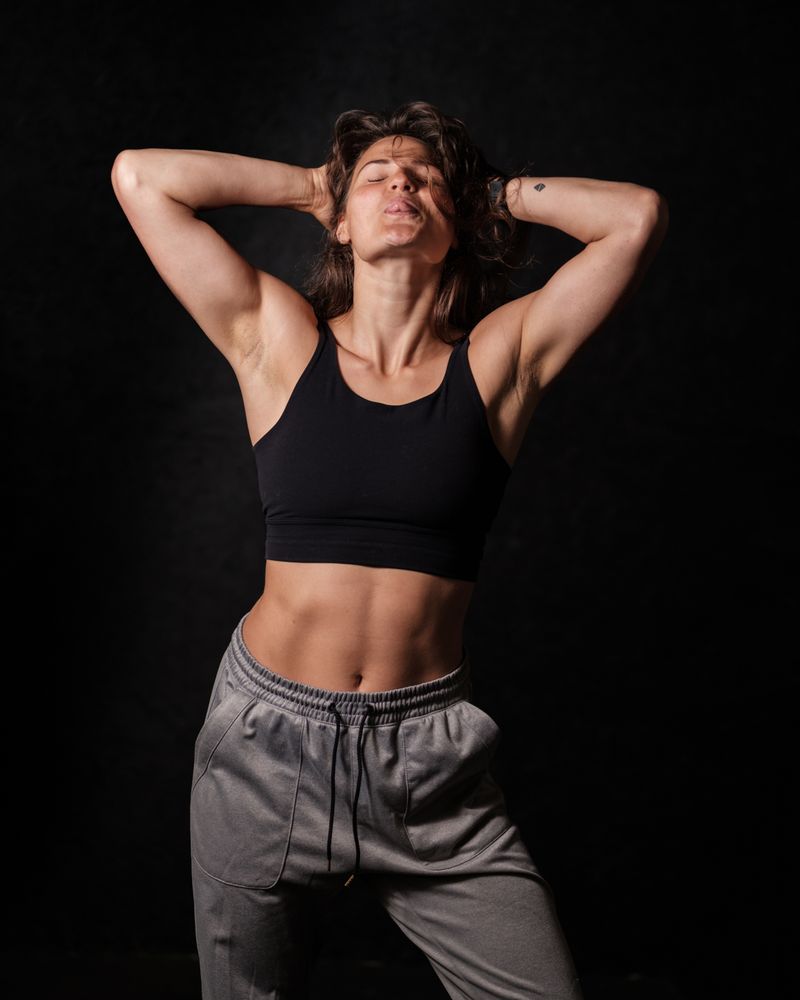 A strong, athletic woman with toned abs poses confidently against a dark background. She wears a black sports bra and grey sweatpants, with her hands resting on her head, eyes closed, and an expression of confidence and empowerment. Photo by Lieven Dewitte