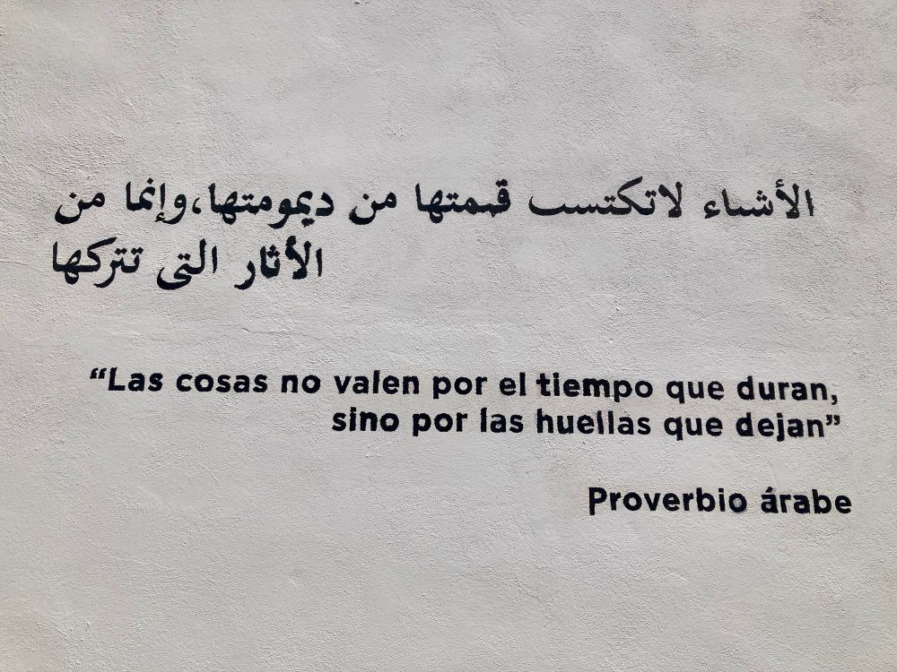 A proverb written on a white wall in Arabic and Spanish. The Spanish text reads: “Las cosas no valen por el tiempo que duran, sino por las huellas que dejan” — Proverbio árabe, which translates to “Things are not valued for how long they last, but for the traces they leave.”