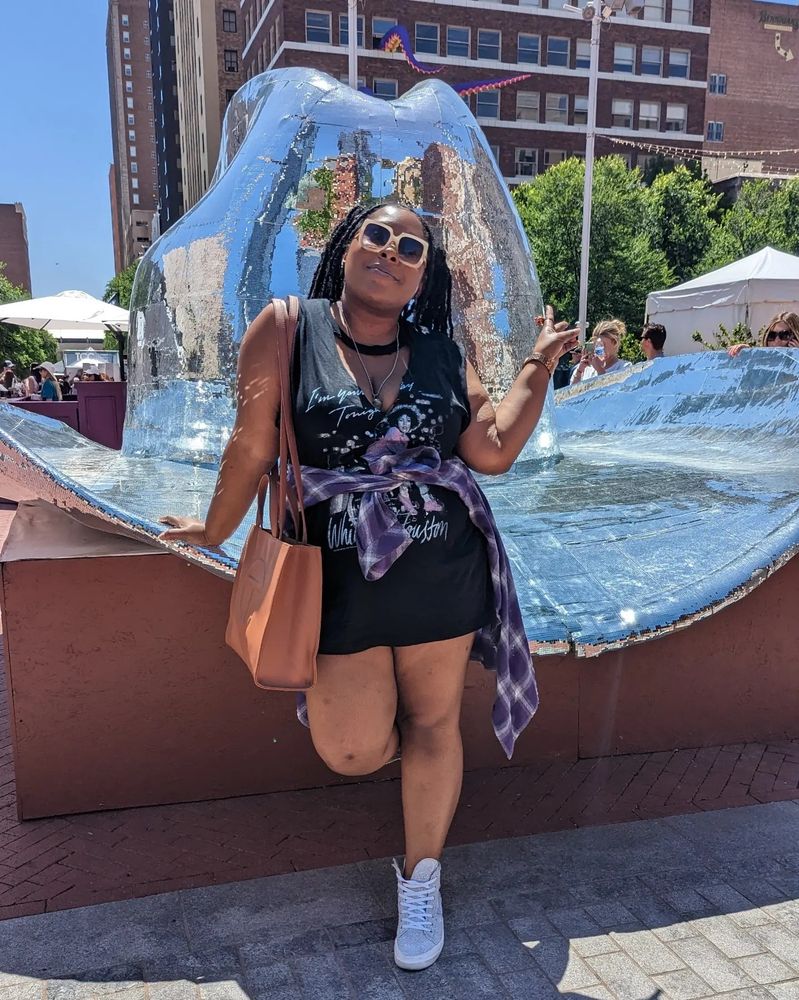 A Black millennial woman in a black dress with a purple jacket tied around her waist, posing in front of a statue of a large silver hat, and to no one's surprise holding up a peace sign. 