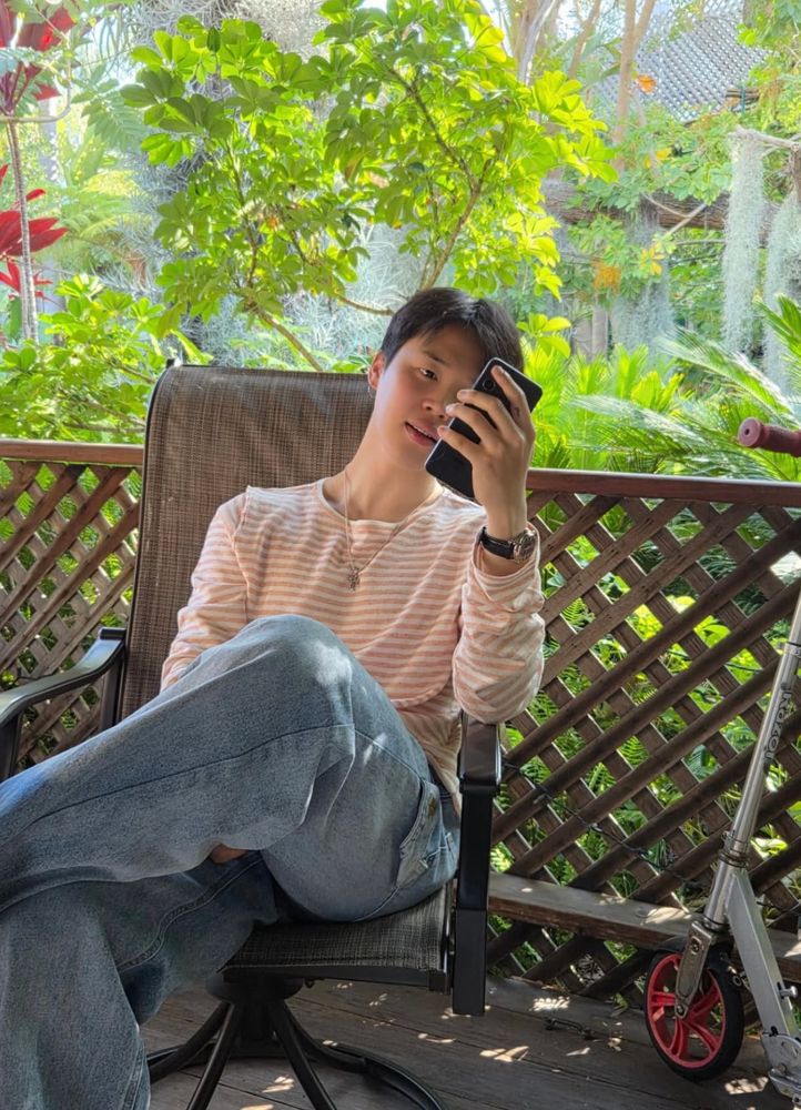 Jimin through Hobi’s lens. Sitting in a chair with a background of trees. He looks ethereal 