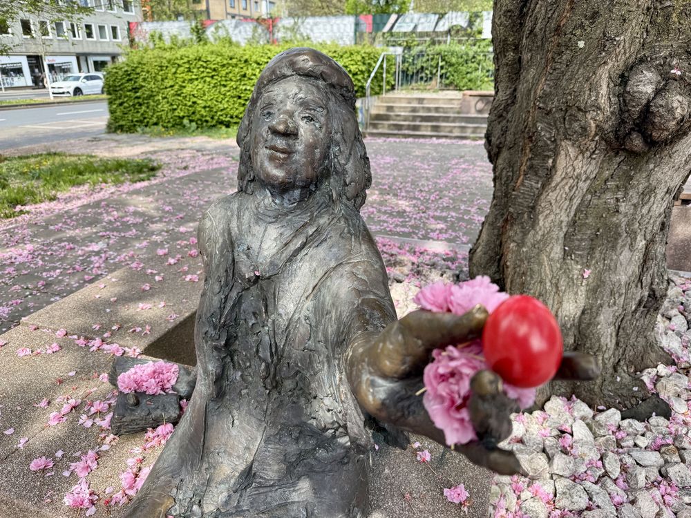 Das Tana-Schanzara-Denkmal im Bochumer Ehrenfeld: Die Bronzestatue der Schauspielerin sitzt unter einem blühenden Baum und hält ein rotes Osterei und rosa Blüten in der Hand. Um sie herum liegen Kirschblüten auf dem Boden.