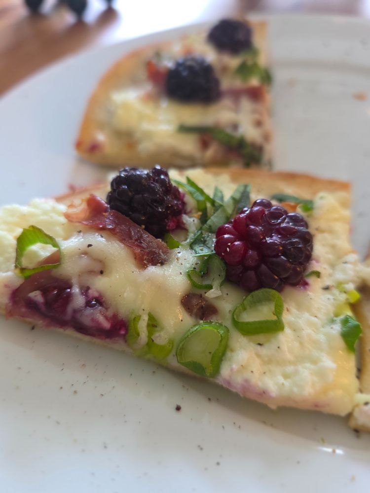 a slice of a pizza with cheese, prosciutto, blackberries, scallions, and fresh basil