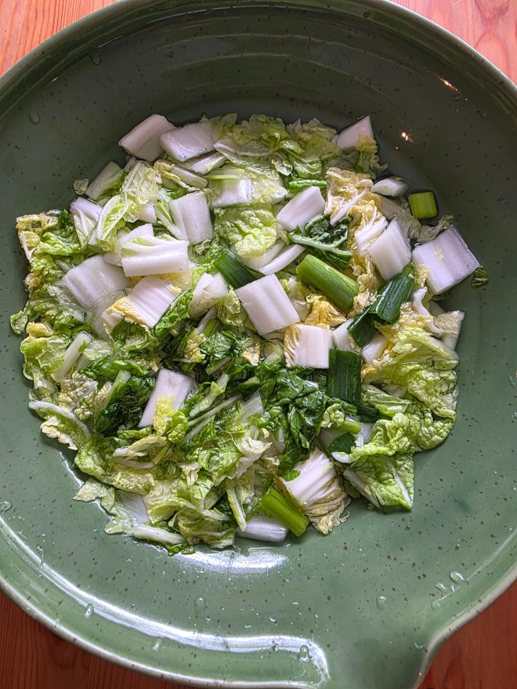 Coarsely chopped napa cabbage and scallions in a strong brine, in a large green bowl on a pine table.