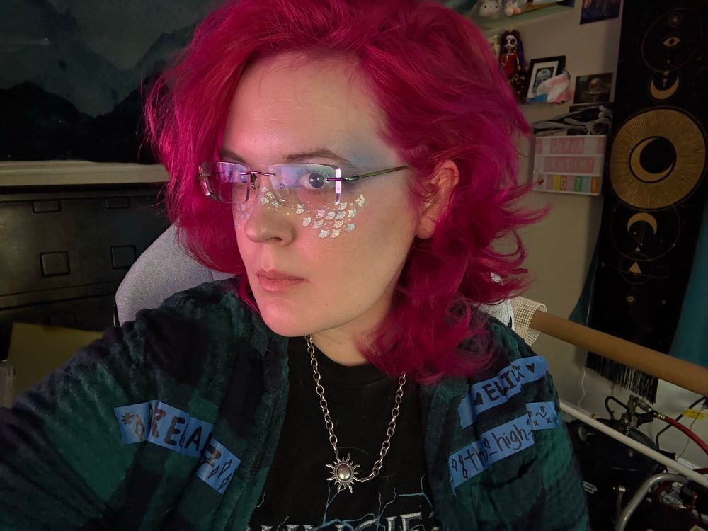 This is a picture of a person with curly, luridly pink hair, and vibrant, iridescent scale makeup. Their glasses are also iridescent. They wear a silver sun charm around their neck, and wear a plaid shirt with strips of painters tape with users names on it. Behind them, there is a lightly cluttered office with many trinkets and Destiny 2 memorabilia. 