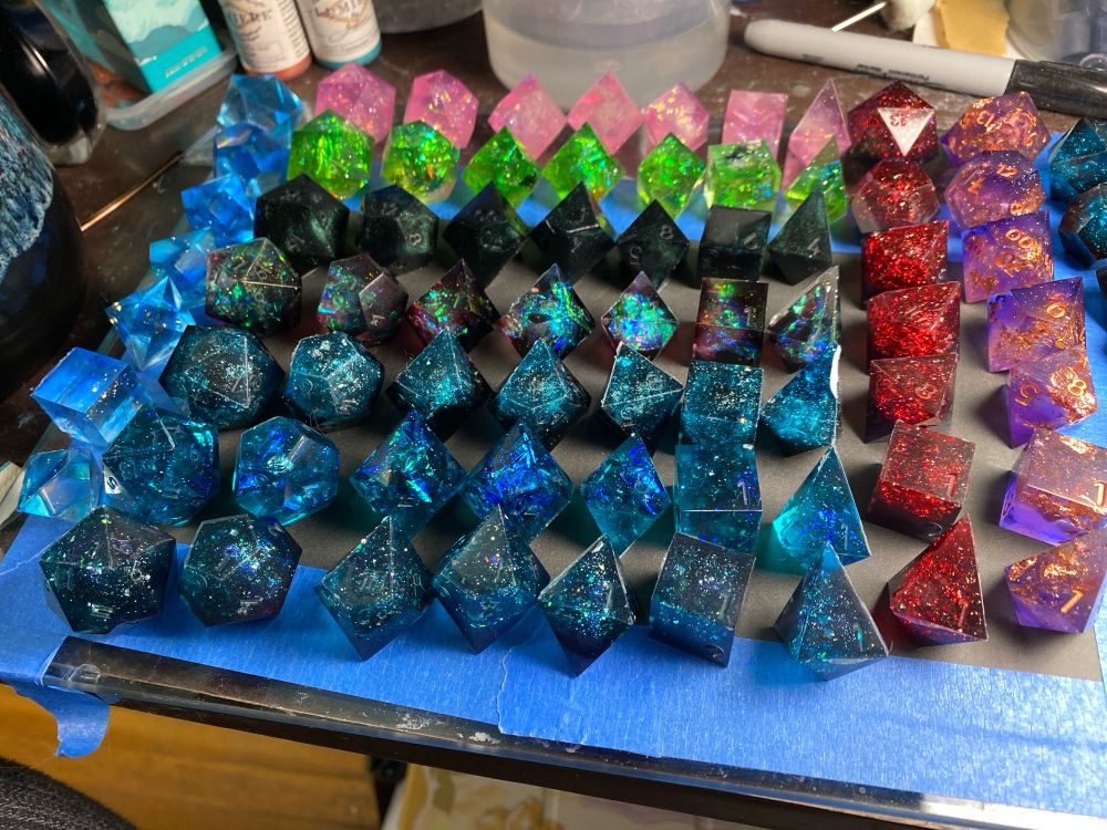 A picture of many, many, many sets of dice. The top row is pink, the second row is green, the third row is opaque black with green interference powder. The fourth row is black red and pale blue with foil. The bottom three rows are black with teal tops and iridescent foil, silver foil and holo flake. A set of pale blue blanks are on the left side of the image. The right side of the image has a set of black and red dice, a set of galaxy-like purple dice with gold foil, and a barely visible set of blanks in black and teal