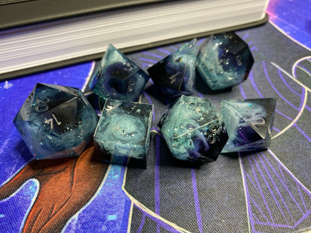  set of cloud-oriented polyhedral dice. They are a black, violet and pale clear blue gradient with pale blue clouds strewn throughout. They have flecks of holographic foil and white mica glitter inside of them. The white clouds are swirled in a spiral to look like mesocyclones. This angle showcases some of the “holes” through the white clouds.