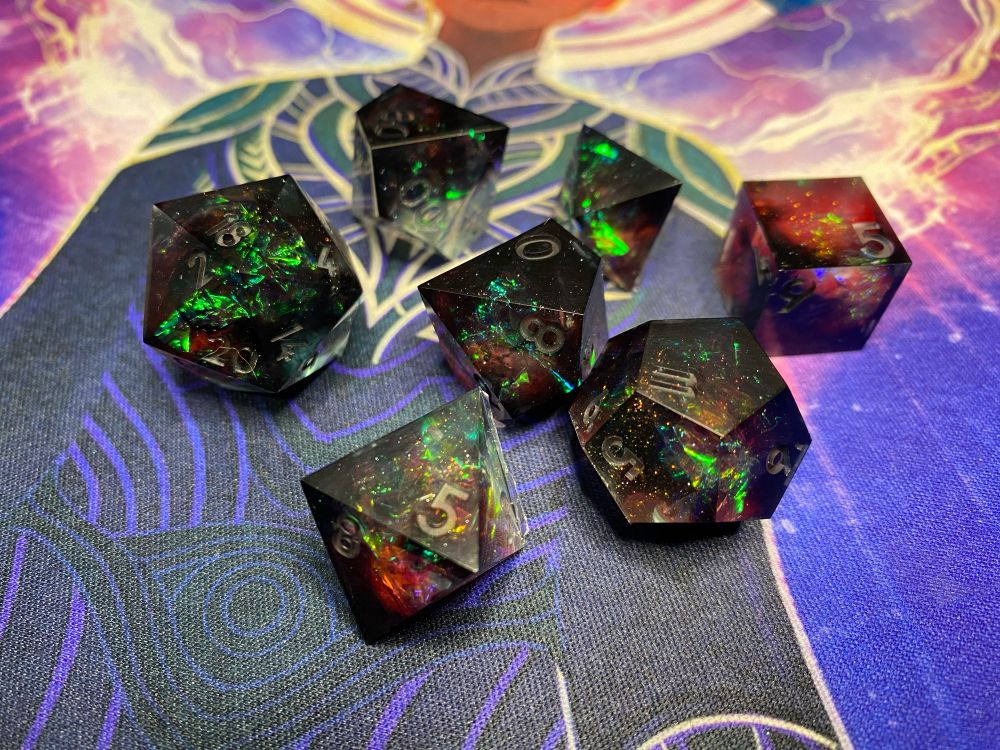 A set of polyhedral dice in black, red and pale blue with green holo foil flashes and hyper holo flake. This is the “bottom” side of the dice and they are more black and red 