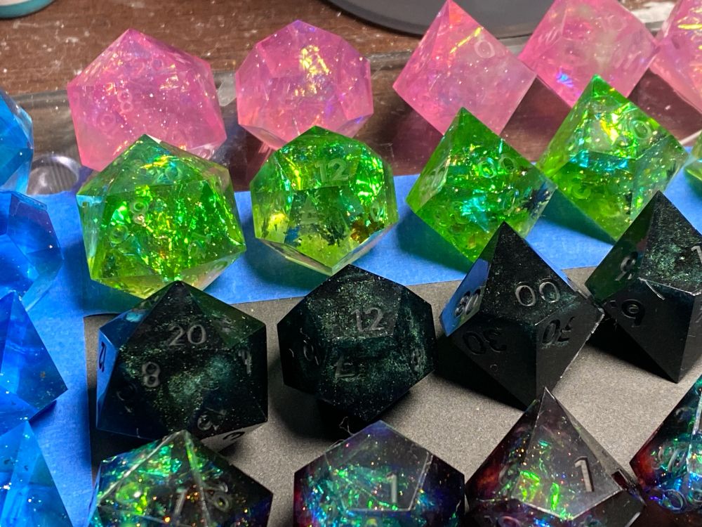A picture of several sets of dice. The top row is iridescent pink with wisps of pale green clouds. The second row is green with iridescent foil. The third row is opaque black with deep green interference powder. The fourth row, barely visible on the bottom is black, red and pale blue with iridescent foil. On the far left of the image, a barely visible set of swirly blue blanks (dice without numbers) sit. 