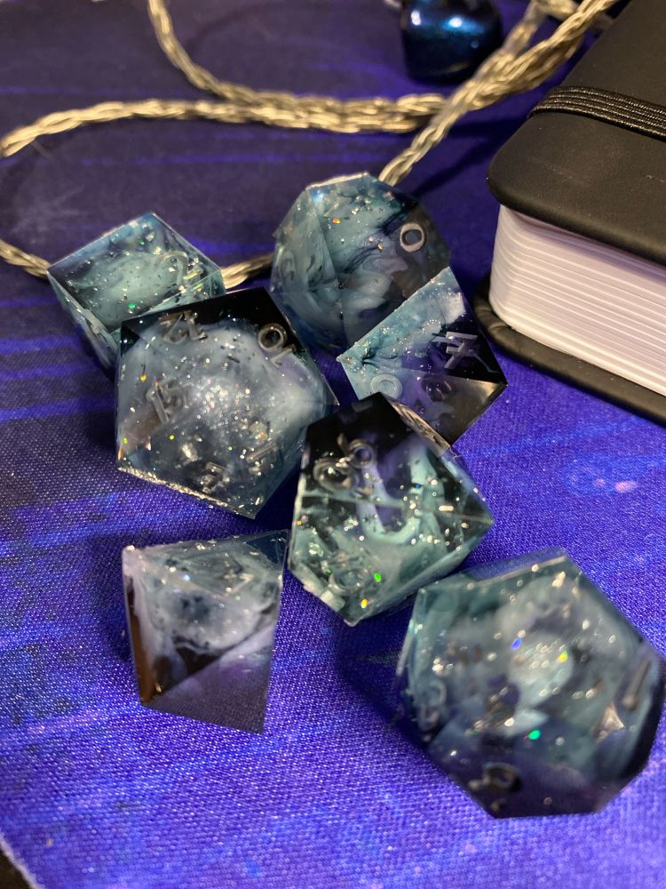 A set of cloud-oriented polyhedral dice. They are a black, violet and pale clear blue gradient with pale blue clouds strewn throughout. They have flecks of holographic foil and white mica glitter inside of them. The white clouds are swirled in a spiral to look like mesocyclones. 