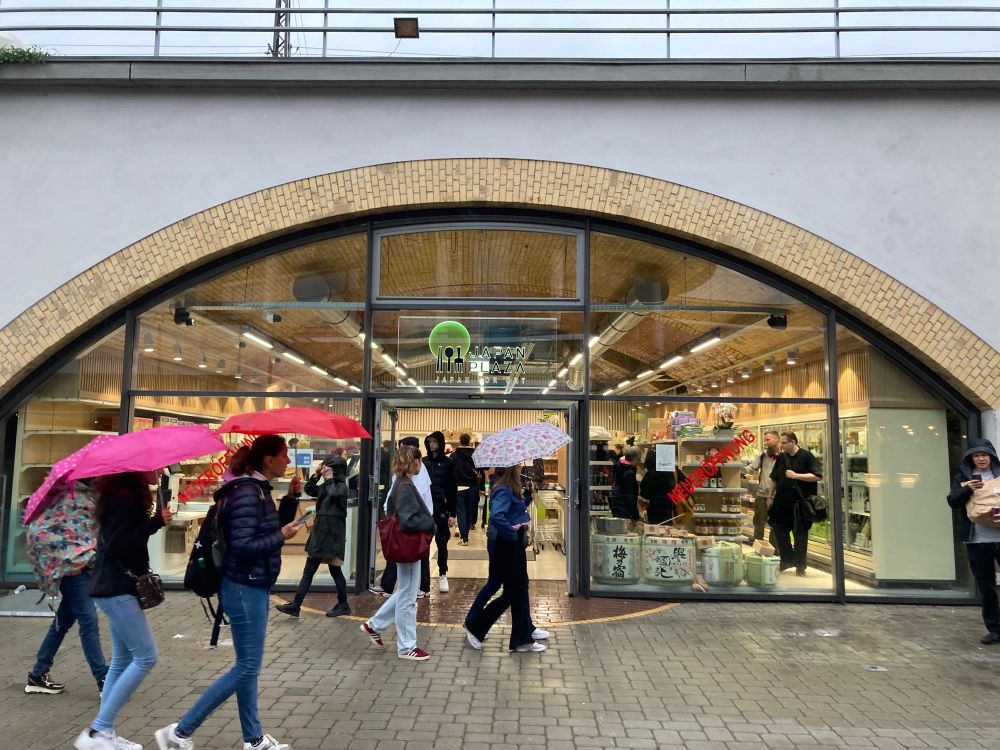 Eingangsbereich des japanischen Supermarktes, im Vordergrund sind Menschen mit Regenschirmen