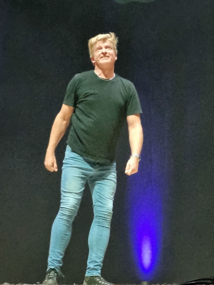 Rhys Darby in skinny blue jeans and black tshirt with a goofy smile and a delightful bit of hair looking very swooshy