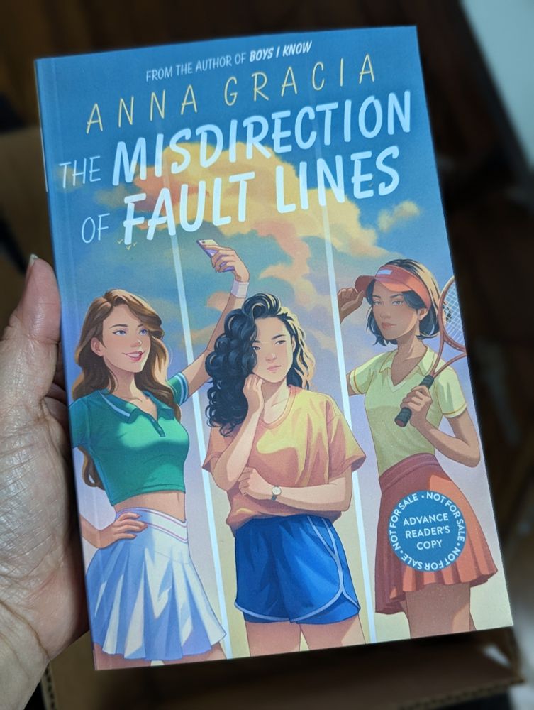 3 girls standing against a turquoise sky with the words THE MISDIRECTION OF FAULT LINES across the top. Girl on left has long brown hair and smiling at her phone for a selfie, girl in middle with curly black hair and a reserved expression, girl on right glaring out with a racquet in hand