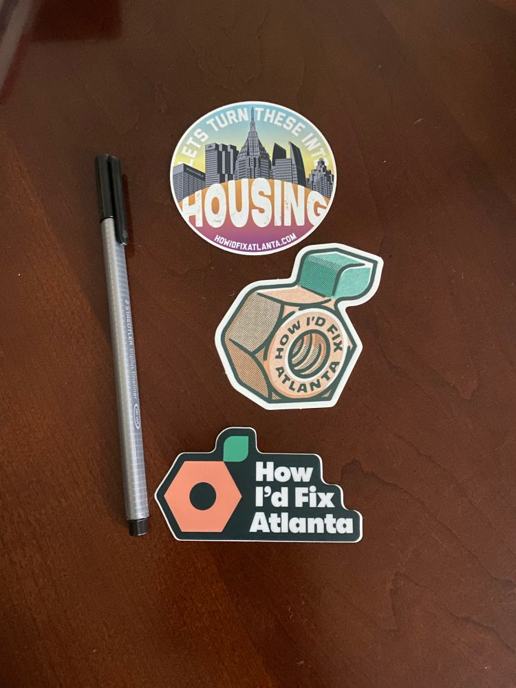 Three stickers next to a pen for size comparison. Top one says “Let’s Turn These Into Housing” with a picture of notable Atlanta skyline office buildings. Middle sticker is of an orange and green Georgia peach stylized as a hex nut with “How I’d Fix Atlanta” around the center. Last sticker is of a similar peach-hex nut logo next to the words “How I’d Fix Atlanta.”