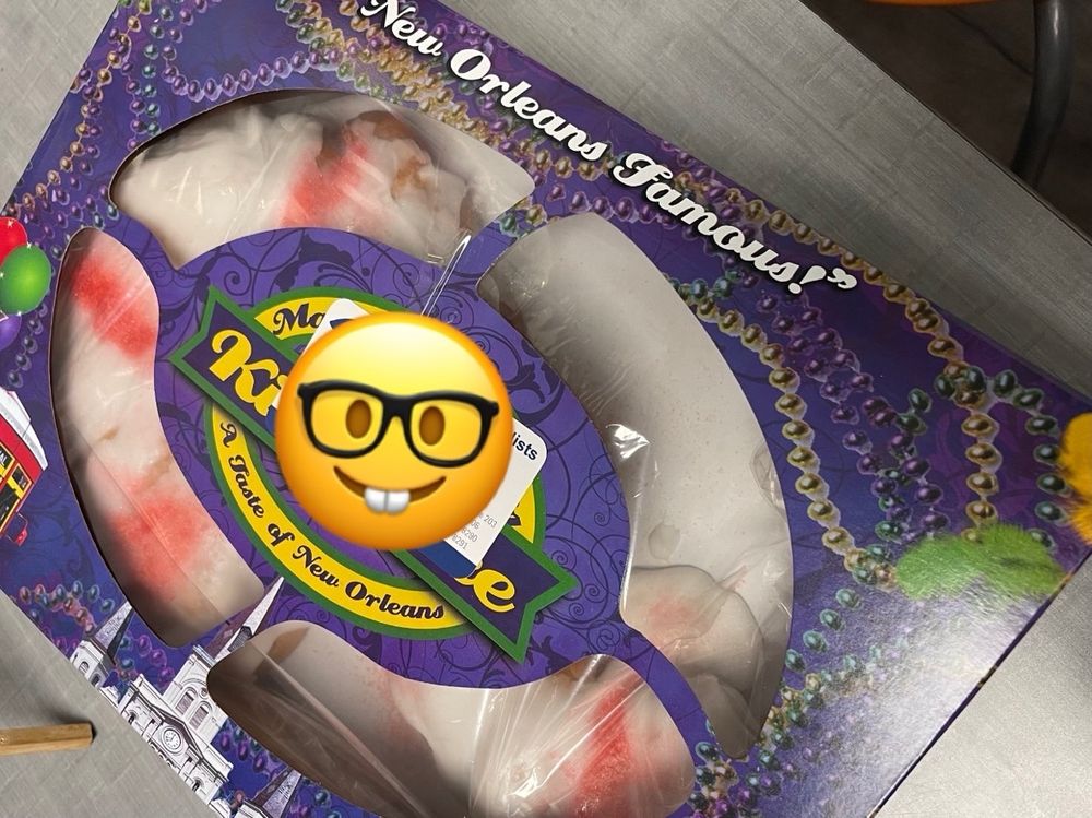 A purple, green, and gold short, wide rectangular box with a clear plastic window showing a king cake that is shaped like a candy cane inside. It’s covered with white sugar icing and disturbing sad looking light red stripes randomly either sprayed on or made with red sugar 