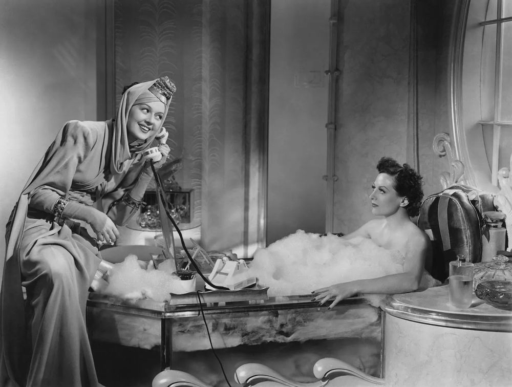 Joan Crawford in a bathtub filled with bubble bath looking annoyed at Rosalind Russel talking on her private telephone