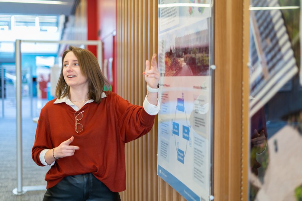 A woman wearing a red jumper points up to a scientific poster on a wooden slat wall.