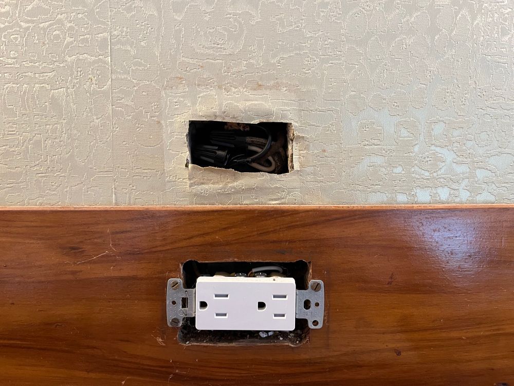 The removal of the patch of wallpaper reveals a hole in the wall and the wires that were spliced to move the outlet down. The wallpaper where the patch sat is much less yellow than the rest