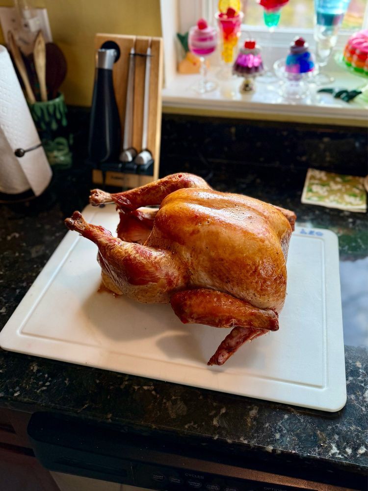 A whole roasted turkey, golden-brown and trussed, resting on a white cutting board on a dark kitchen countertop. The background shows the kitchen window decorated with colorful plastic or glass dessert novelties, a paper towel holder, and a knife block.