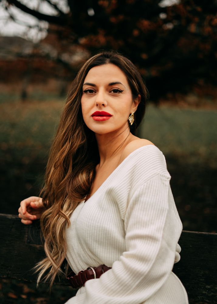 Model posing in bench looking towards camera with dark brown hair, red lips, olive skin. Wearing a white dress 