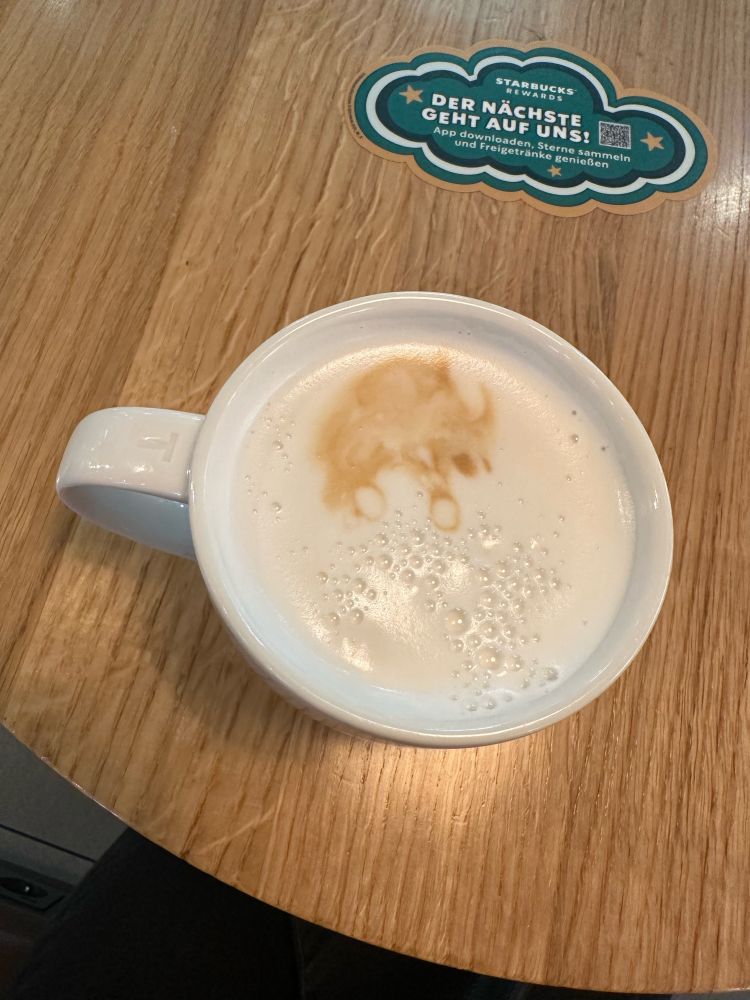 Tasse von oben fotografiert. Zu sehen ist die Crema, deren Muster für mich wie ein Nashorn auf Baumstämmen (im Querschnitt) oder auf Bällen aussieht. 
Die Tasse steht auf einem Holztisch, auf dessen Mitte ein Aufkleber prangt (Werbung für Starbucks).