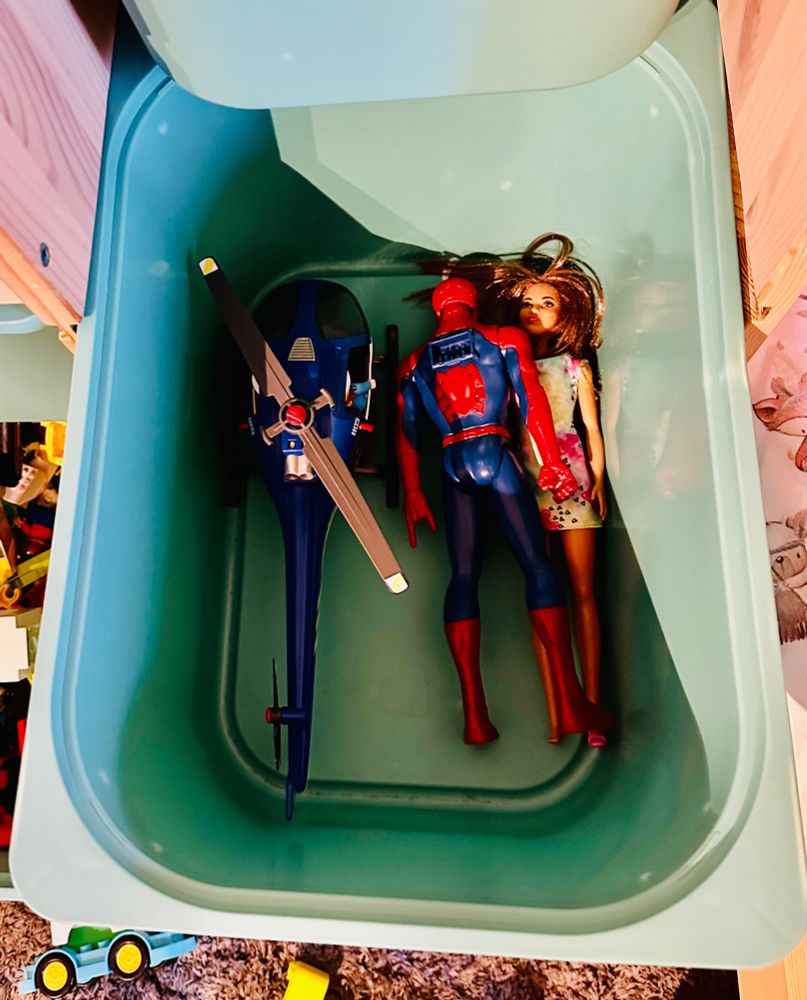 Gesuchte Barbiepuppe liegt halb unter einer Spider-Man-Figur in einer Kiste. Daneben ist ein Playmobil-Hubschrauber.