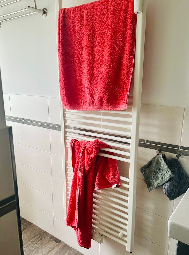 In einem zur Hälfte gefliesten Raum ist ein Handtuchheizkörper. Über den oberen Bereich ist ein Handtuch halbwegs ordentlich drapiert, im unteren Bereich ist ein Handtuch zwischen die Streben gewurschtelt. Beide Handtücher sind korallrot. Rechts neben dem Heizkörper hängen noch zwei graue Waschlappen. 