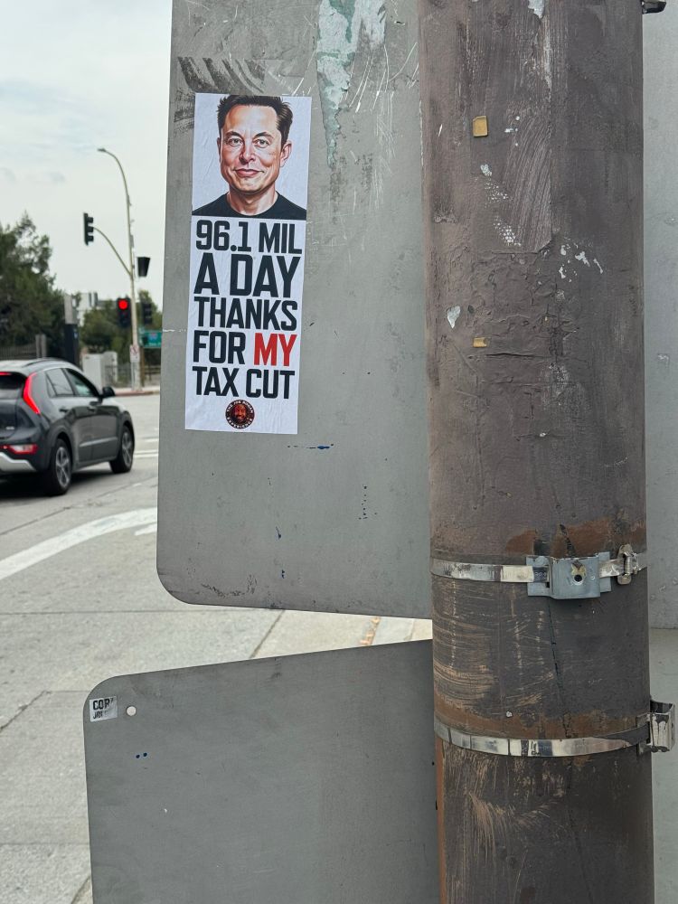 Sticker on the back of a metal street sign reads: “96.1 MIL A DAY THANKS FOR MY TAX CUT” with a picture of a smiling Elon Musk and a Joe Rogan Experience logo at the bottom.