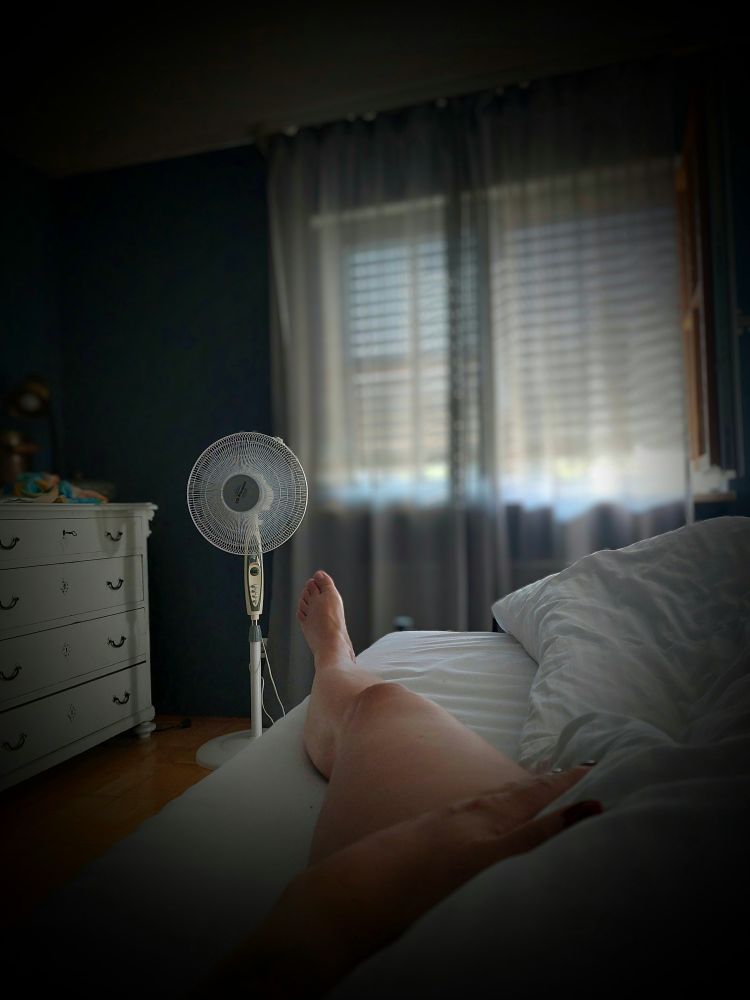 Ein Bein schaut unter der Bettdecke hervor, im Hintergrund ein Ventilator 