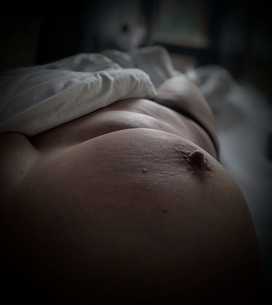 Weibliche rechte Körperhälfte, von der Schulter abwärts im Bett liegend fotografiert, die linke Hälfte unter der Decke, die Brustwarze zur Knospe zusammengezogen, sanftes Morgenlicht fällt auf den Körper 
