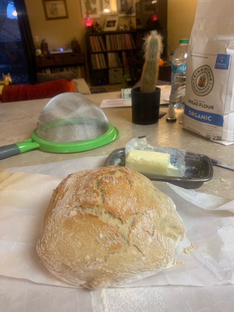 A golden brown, round, rustic loaf of bread sits on parchment paper next to some butter and a bag of flour 