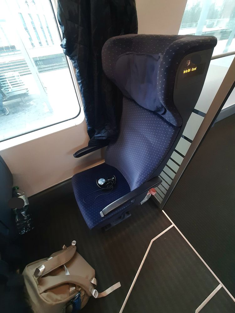 Ein einzelner Sitz mit dunkelblauem Polster im ICE, direkt am Durchgang zum Fahrradbereich.
Auf dem Sitz liegen schwarze Kopfhörer, davor ein beiger Rucksack und im Netz des Vordersitzes befindet sich eine große Wasserflasche.
