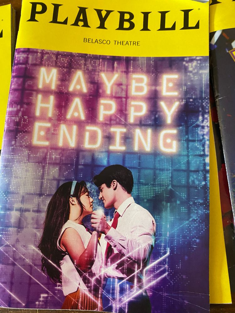 Playbill program for Maybe Happy Ending. The background is blue and purple and pink with circuit board lines. The title is in a futuristic font. The two lead characters are on the front. One is an Asian woman with black hair  and blue headband, white top and hello skirt. The other is a man with black hair, red tie, white shirt, and blue pants.