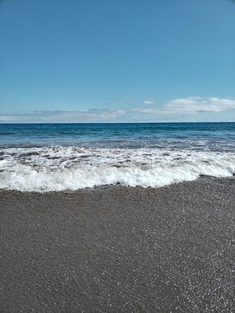 L'oceano a Maspalomas (Gran Canaria, Spagna): le onde spumose si infrangono sulla sabbia. Il cielo è azzurro, c'è qualche nuvoletta bianca. 