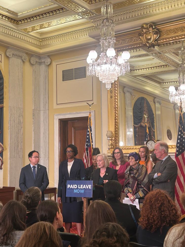 Senator Gillibrand surrounded by members of Congress at a podium that says “paid leave now”