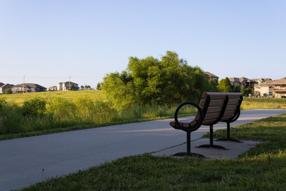 A photo of a walking path at sunset with houses and trees  in the background and a park bench in the foreground.
