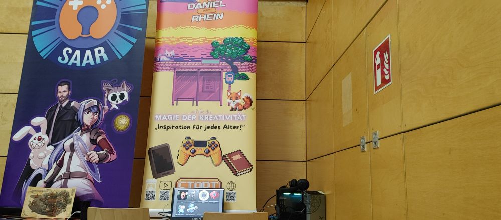 Stand auf der Retrocon 2025 8.11 - 9.11.2025. auf dem Bild ist ein Banner von Daniel Rhein Gamedev zu sehen. Rechner, Stühle und Controller stehen bereit für die Besucher. 