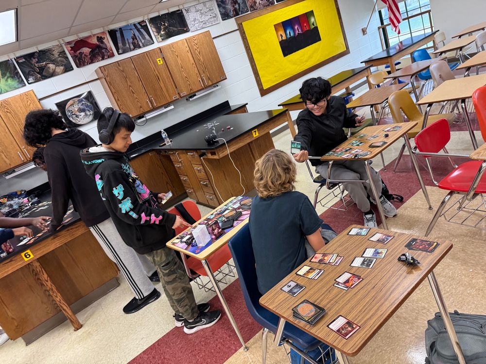 3 students in highschool desk-chair furniture all facing the same direction, they’re rather awkwardly leaning over to see each others’ cards.